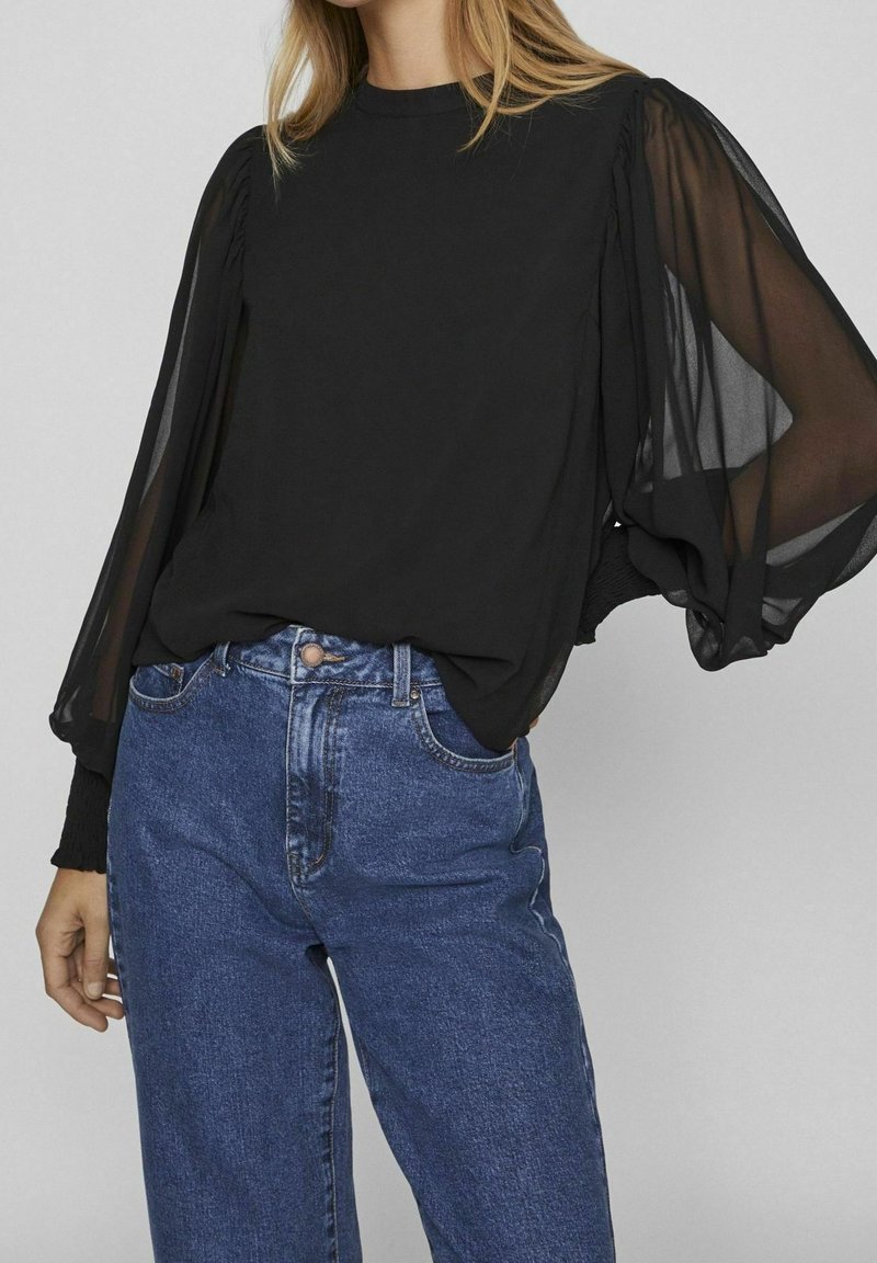 Blusa nera con maniche a sbuffo trasparenti, collo rotondo e polsini elasticizzati. Indossata con jeans in denim azzurri a vita alta. Tessuto liscio in tutto.