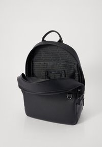 Sac à dos en cuir noir avec double fermeture éclair, sommet arrondi, poche avant et un intérieur présentant un motif de logo répétitif. Texture lisse.