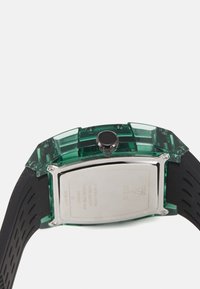 Caja de reloj de plástico verde transparente con un fondo metálico plateado, correa de goma negra y una corona circular en el lateral.