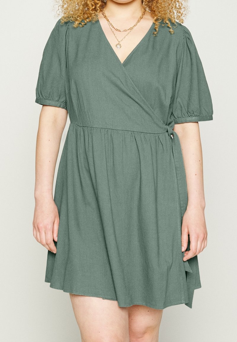 Femme portant une robe portefeuille verte à manches courtes avec une ceinture nouée à la taille et deux colliers en or superposés.
