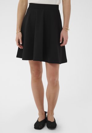 Femme portant une jupe noire mi-longue, un haut blanc, des ballerines noires et des bracelets en or, debout devant un fond uni.