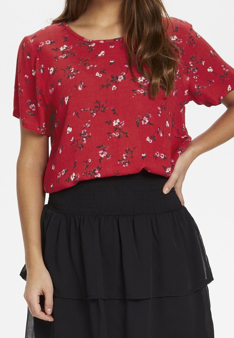 Femme portant un chemisier rouge à fleurs à manches courtes, rentré dans une jupe noire à volants, une main sur la hanche et l'autre pendant le long du corps.