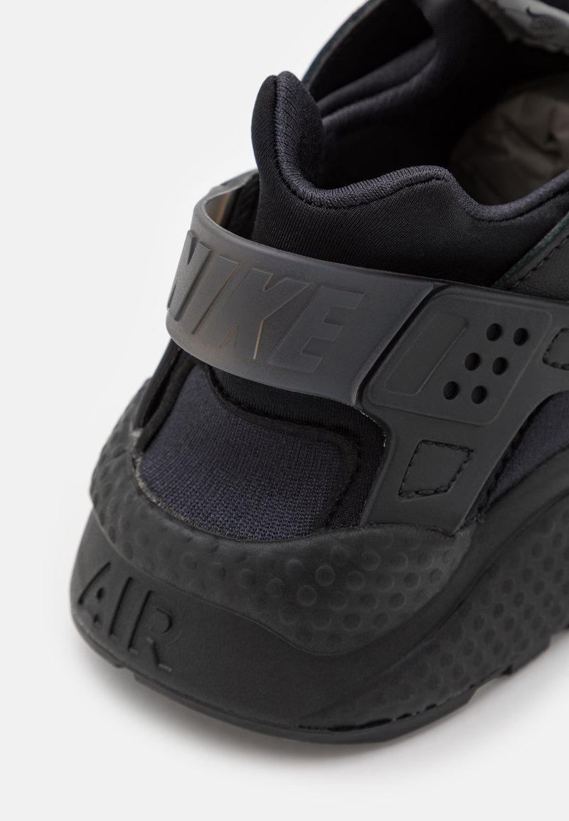 Schwarzer Sportschuh mit strukturiertem Gummisohle, gepolstertem Knöchelkragen und verstellbarem Riemen mit eingeprägtem Branding. Glatte Stoffakzente.