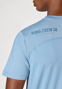 Hele sinine lühikeste varrukatega ümmarguse kaelusega T-särk, millel on siledast kangast valmistatud, ning selja õlal on tumehallis trükitud tekst "PUMA CREW '48".
