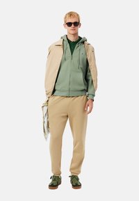 Sudadera con capucha verde con cremallera, combinada con una chaqueta beige, pantalones deportivos beige y zapatillas verdes. Los accesorios incluyen gafas de sol negras.