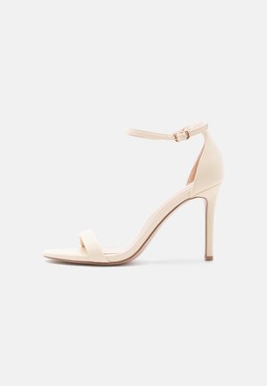 Beežid kõrge kontsaga sandaalid pahkluu rihmaga ja avatud varbaga, sile viimistlus ning kitsas konts valgel taustal.