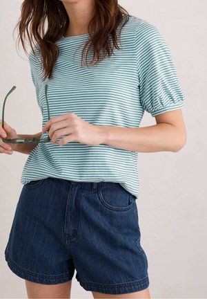 Gestreiftes türkis-weißes Oberteil mit kurzen Puffärmeln, kombiniert mit dunklen Jeansshorts. Das Model hält eine grüne Sonnenbrille vor sich.