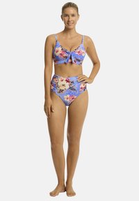 Modella che sorride in un bikini con motivo floreale, in piedi a piedi nudi contro uno sfondo neutro, che mette in mostra uno stile estivo da bagno.