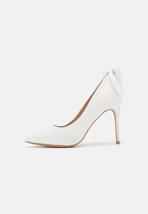 Witte pump met hoge hak en puntige neus, gemaakt van glad materiaal, met een fijn strikdetail bij de hak.