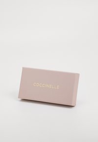 Boîte de marque Coccinelle de couleur rose doux, de forme rectangulaire, avec des lettres dorées, texture mate lisse, bords nets et design minimaliste.