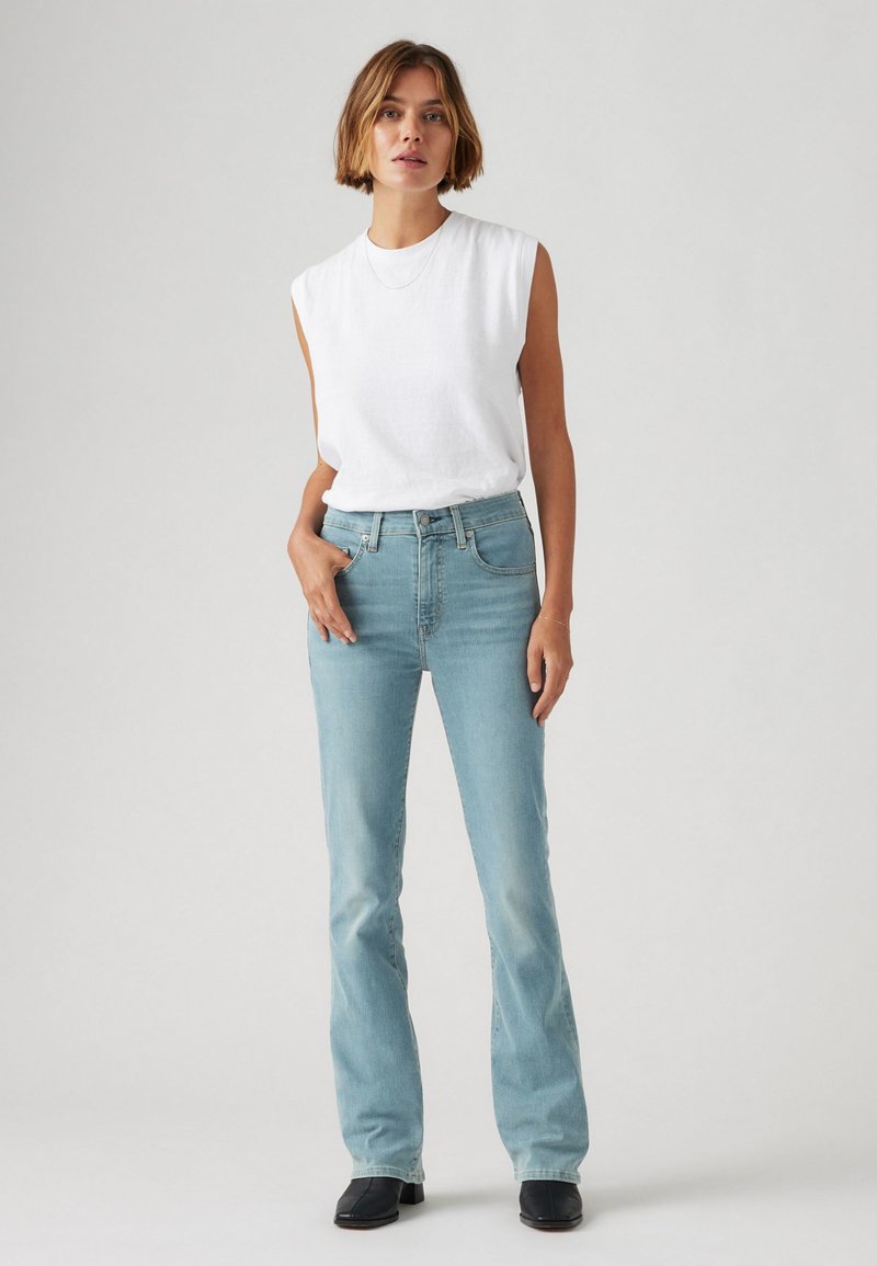 Jeans de campana azul claro con cintura alta, combinados con una blusa sin mangas blanca. Los jeans presentan una textura de denim clásica y un leve desgastado.