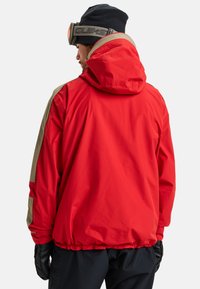 Man in een rode jas met beige strepen op de mouwen, zwarte handschoenen, marineblauwe broek, zwarte muts en skibril, zijwaarts gericht op een witte achtergrond.