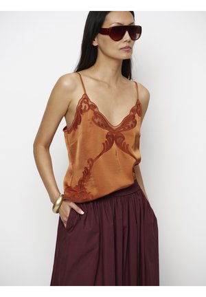 Vrouw met grote bruine zonnebril, oranje satijnen hemdje met kantdetail, bordeaux rok en gouden armband met één hand in de zak.