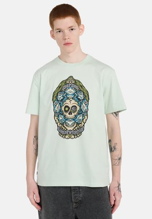 Junger Mann trägt ein hellgrünes T-Shirt mit einem bunten grafischen Totenkopf und Tiergesichtern, umgeben von den Texten "Seekers" und "Element & Timber".