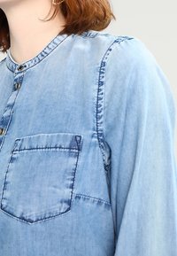 Camicia in denim blu chiaro con scollatura rotonda, tasca frontale e dettagli con bottoni. Il tessuto è morbido con cuciture visibili e una texture sbiadita.