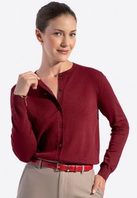 Cardigan rosso con scollatura rotonda, maniche lunghe e chiusura a bottoni. Indossato con una cintura color crema, caratterizzata da una texture liscia e un design aderente.