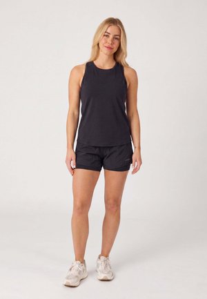 Frau trägt ein schwarzes ärmelloses Top, schwarze Shorts und weiße Turnschuhe und steht vor einem schlichten hellen Hintergrund.