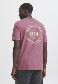 Lilla bomulds t-shirt med rund beige grafik tekst på ryggen. Har korte ærmer og en afslappet pasform. Simpelt design, ingen synlige mønstre.