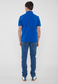 Maglietta polo blu con colletto, abbinata a pantaloni jeans blu aderenti. La maglietta è di un colore uniforme, mentre i jeans presentano tasche posteriori con dettagli di cucitura.