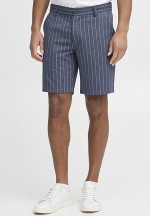 Shorts pour hommes en bleu marine, fabriqués dans un tissu texturé avec des rayures verticales blanches, dotés d'une fermeture à bouton et de passants de ceinture. Portés avec des baskets blanches.