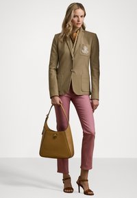 Beige-viimeistely takki, jossa on taskuyksityiskohta, yhdistettynä vaaleanpunaisiin farkkuihin. Pitelee ruskeaa hobo-kassia; ruskeat remmisaappaat täydentävät asua.