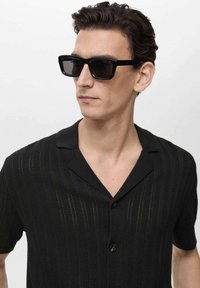 Black short-sleeve shirt with a textured pattern and a V-neck collar. The model wears black square sunglasses.