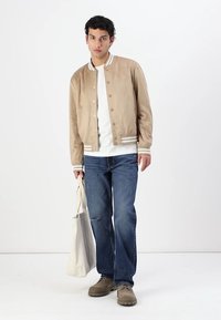 Beige semsket bomberjakke med hvit kant, båret over en hvit T-skjorte, kombinert med blå jeans og brune sko. Modell som holder en beige tote bag.