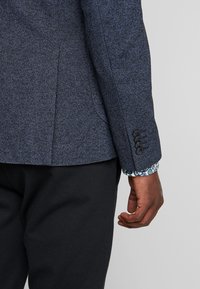 Tmavě modrý texturovaný blazer se štíhlým střihem, s dvojitým rozparkem, černými knoflíky a vzorovými detaily na manžetách. Tkanina odolná proti pomačkání.