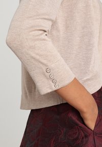 Beige knit sweater with three-quarter sleeves, featuring four light buttons on the cuff. Smooth texture and fitted design, paired with a dark patterned skirt.