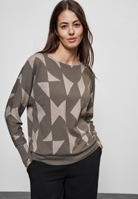 Pull gris-vert avec des motifs géométriques beiges, col rond et manches longues. Porté avec un pantalon sombre, présenté sur fond uni.