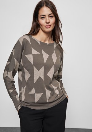 Pull gris-vert avec des motifs géométriques beiges, col rond et manches longues. Porté avec un pantalon sombre, présenté sur fond uni.