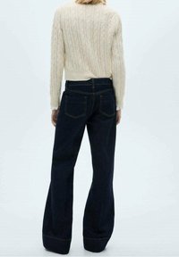 Suéter de punto color crema con un patrón texturizado combinado con vaqueros acampanados azul oscuro que presentan bolsillos traseros y detalles de costuras en contraste.