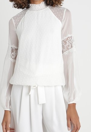 Biała przezroczysta bluzka z kropkowaną strukturą, koronkowymi wstawkami na długich rękawach, wysokim, falbankowym kołnierzem, w połączeniu z białymi spodniami z wiązanym w pasie paskiem.