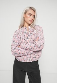 Pletený kardigan v pastelově růžové barvě s barevnými skvrnami. Má kulatý výstřih, zapínání na knoflíky a žebrované manžety a zakončení. Měkká textura.