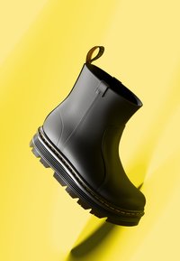 Botín de cuero negro con suela gruesa, dibujo de ondas en la suela, costuras amarillas y una pestaña para tirar con un logotipo. Colocado sobre un fondo amarillo.