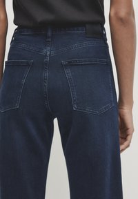 Jeans in denim blu scuro con una trama liscia, caratterizzati da una vita a media altezza, due tasche posteriori e dettagli di cucitura minimalisti.