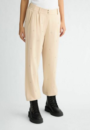 Pantalons beiges avec une coupe décontractée, présentant des motifs d'yeux brodés, des jambes larges et une taille texturée. Associés à des bottes de combat noires.