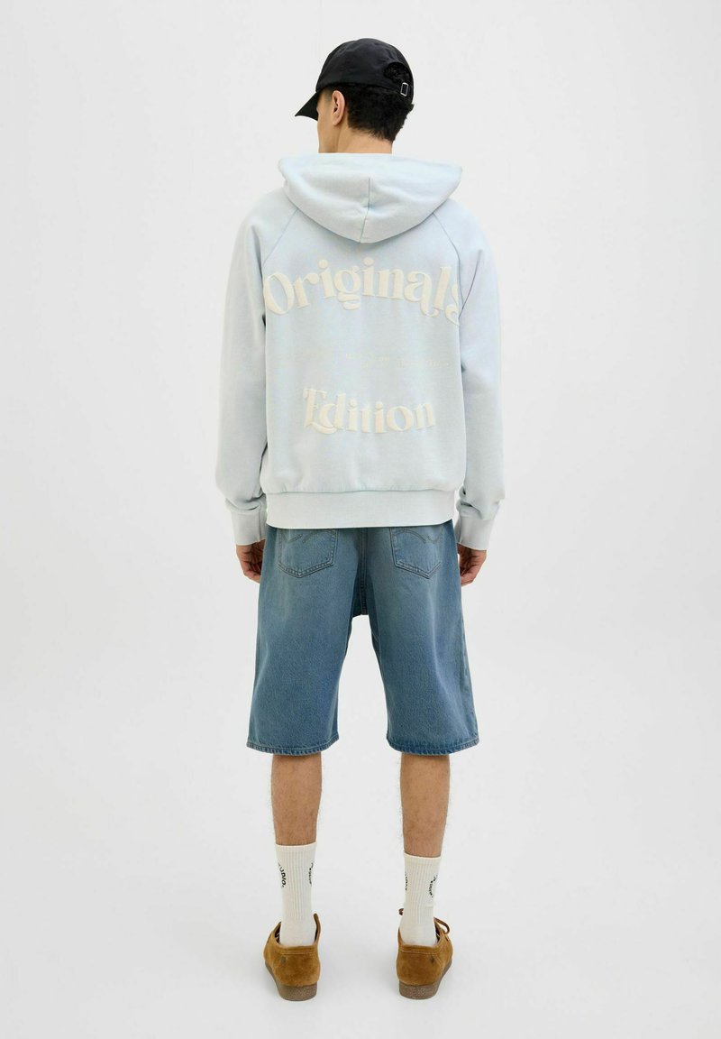 Light blue hoodie with text “Original Edition” in beige on the back, paired with blue denim shorts and tan shoes, white socks.