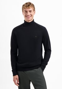 Zwarte coltrui met een gladde textuur en geribbelde manchetten, voorzien van een klein hexagonaal logo op de borst. Model draagt donkere broek.