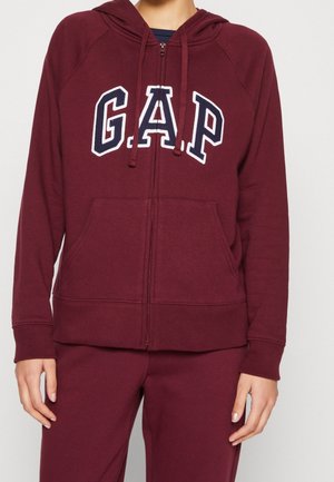 Persoon draagt een bordeauxrode GAP-hoodie met rits en voorkantzakken, gecombineerd met bijpassende bordeauxrode broek, tegen een effen achtergrond.