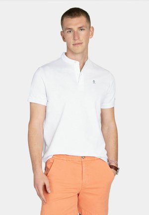 Młody mężczyzna ubrany w białą koszulkę polo z krótkim rękawem i pomarańczowe spodenki, stoi z jedną ręką w kieszeni na jednolitym jasnym tle.