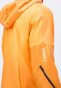 Jachetă portocalie strălucitoare cu glugă, având textul „TERREX” pe mânecă și un detaliu cu dungă diagonală neagră.