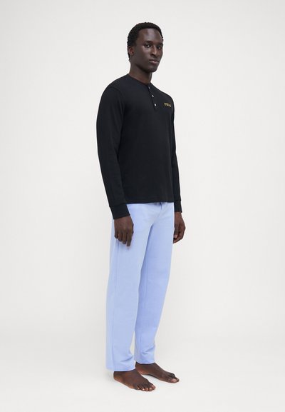 Camisa henley de manga longa preta com decote botão e logotipo dourado, combinada com calças de perna reta azul claro. Tecido suave, ajuste casual.