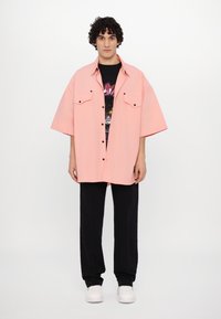 Camisa rosa de gran tamaño con dos bolsillos en el pecho, botones negros, superpuesta a una camiseta gráfica negra, combinada con pantalones negros y zapatillas blancas.