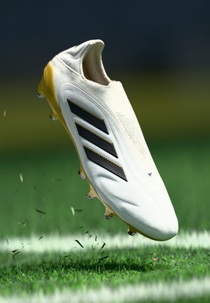 Weiße Fußballschuhe mit einer gelben Sohle, schwarzen Akzentstreifen, strukturierter Oberseite und sichtbaren Nieten, über grünem Gras schwebend.