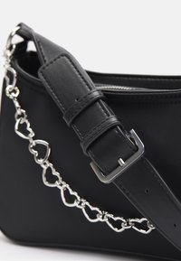 Sac à main noir avec bandoulière en cuir réglable et chaîne argentée ornée de maillons en forme de cœur interconnectés.