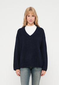 Sötétkék, oversized kötött pulóver mély V-nyakkal és bordás mandzsettával, fehér, magas nyakú felsőre húzva, világoskék farmerral párosítva.