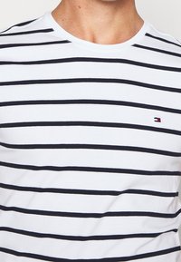 Mies, joka pitää valkoista pyöreäkaulaista t-paitaa, jossa on mustia vaakasuoria raitoja ja pieni punainen, valkoinen ja sininen logo vasemmassa rinnassa.