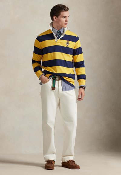 Μακρυμάνικο κοντοζυγωμένο πουκάμισο με κίτρινες και ναυτικές ρίγες, λευκό παντελόνι, καφέ παπούτσια και πολύχρωμη ζώνη. Φορεμένο πάνω από μπλε και άσπρο ριγωτό πουκάμισο.