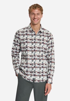 Glunderende man met een geruit overhemd met lange mouwen met abstracte patronen en donkere broek, staand met één hand in de zak.
